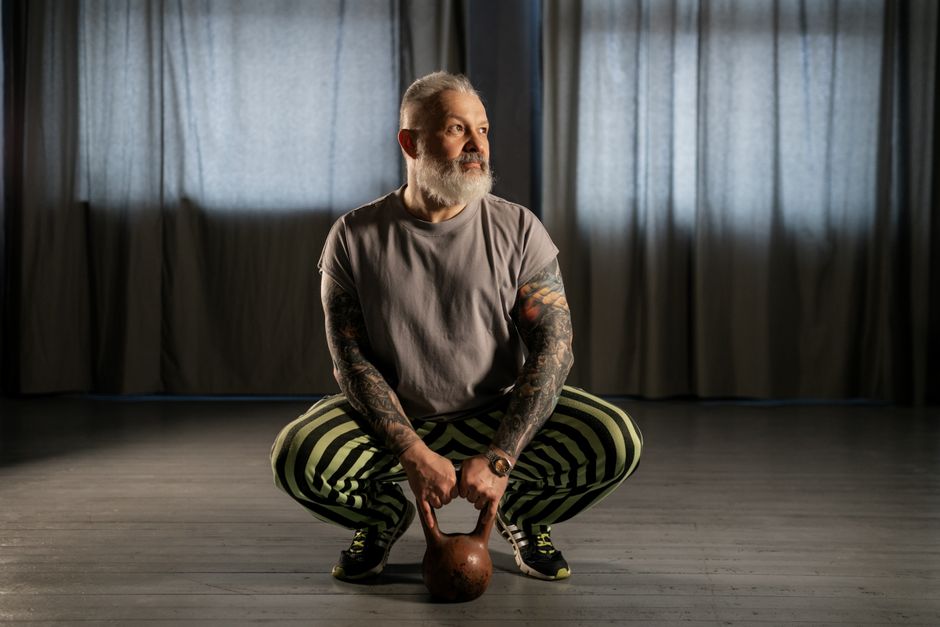 Um homem idoso em agachamento, com as pernas inclinadas e os braços apoiados no chão, demonstrando força e equilíbrio