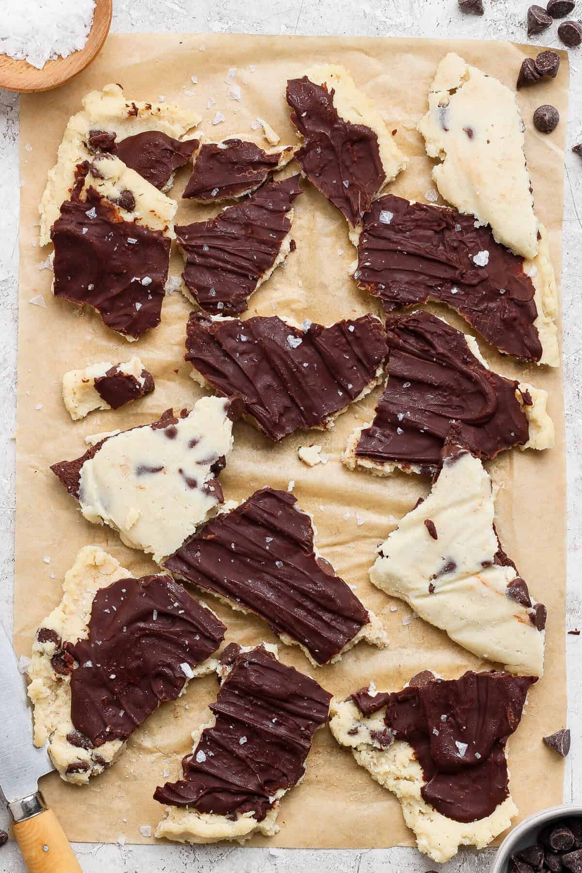 Mãos segurando massa de cookie dough branca com pedaços de cottage cheese, farinha e colher de baunilha sobre bancada