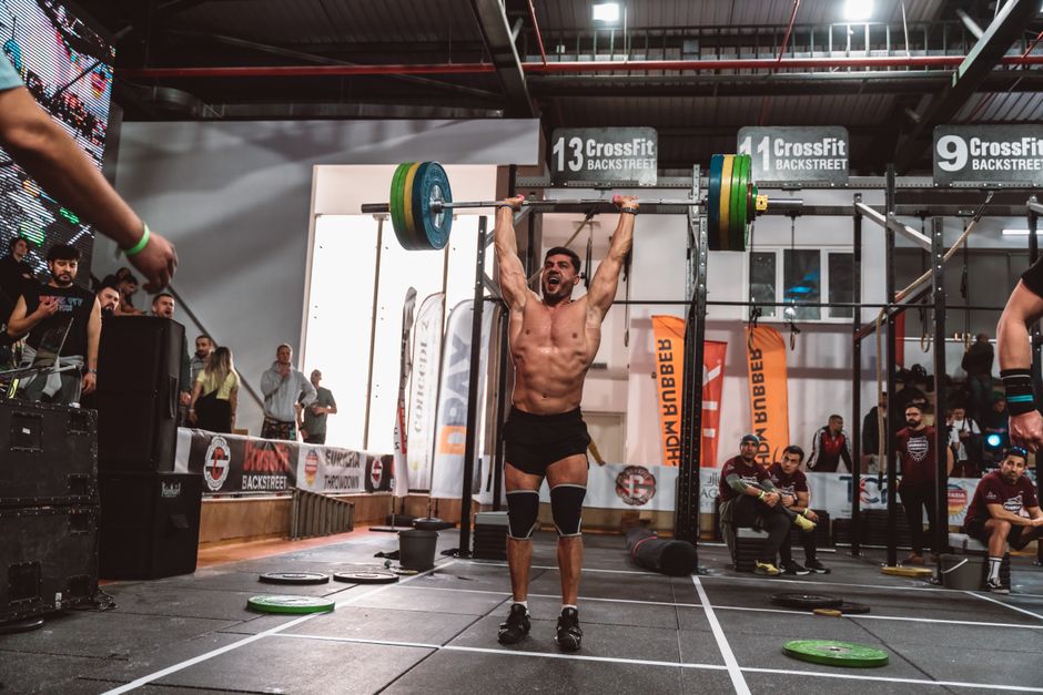Atletas masculinos e femininos correndo em uma trilha de obstáculos, com remos e halteres ao fundo