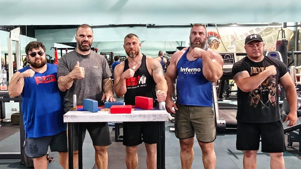 Um atleta fortemente musculado, com braços de grande circunferência, segurando uma barra de armwrestling com expressão de concentração