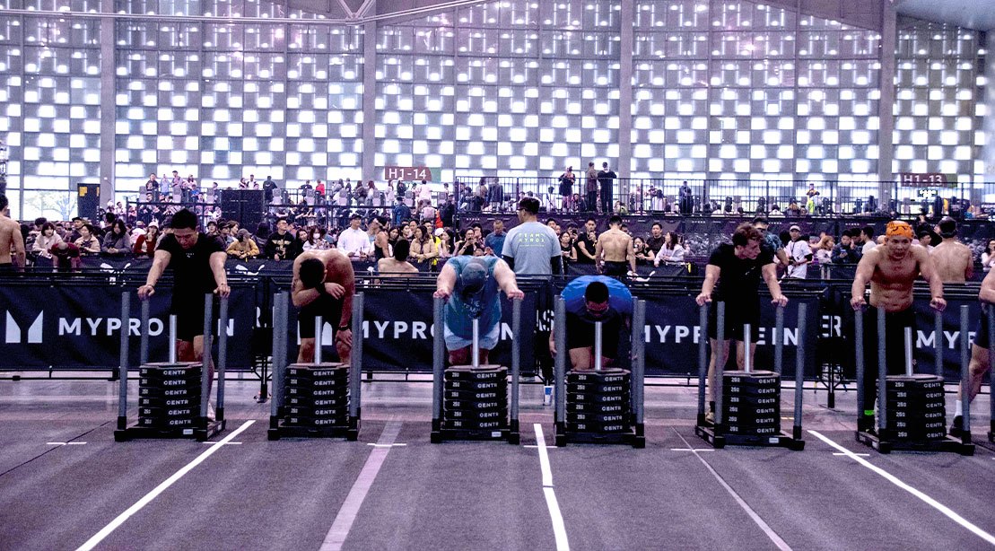 Um atleta em movimento, com um copo de água em sua mão, enquanto faz um exercício de força em um ginásio