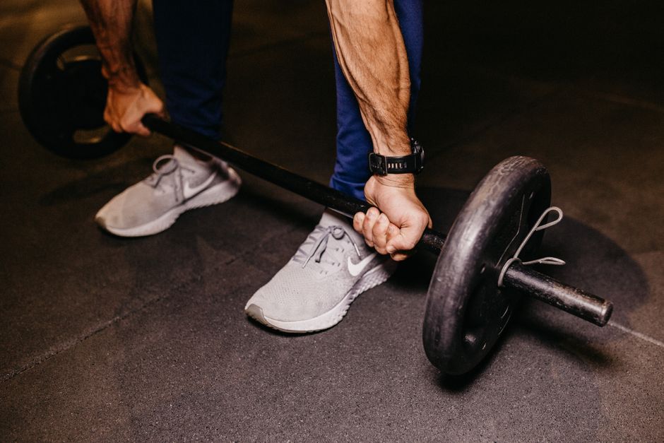 Atleta em pé, vestindo shorts de compressão, agachando com barra carregada enquanto ao fundo há kettlebells e um cronômetro