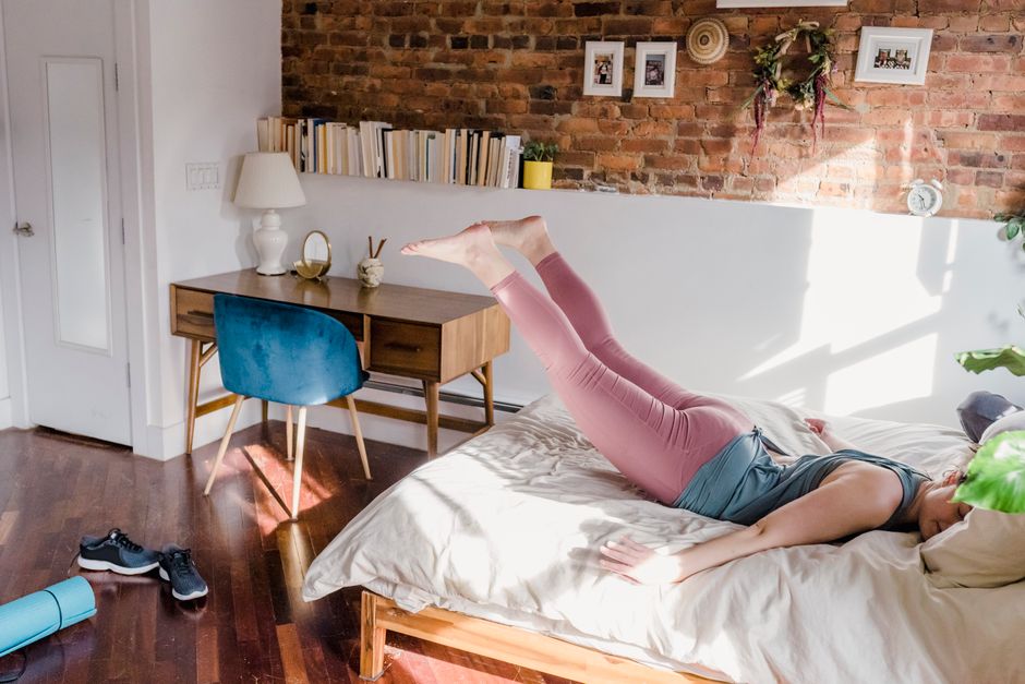 Mulher estendida na cama, com pernas flexionadas e braços segurando pesas ou garrafas, realizando exercícios para fortalecer glúteos e quadríceps