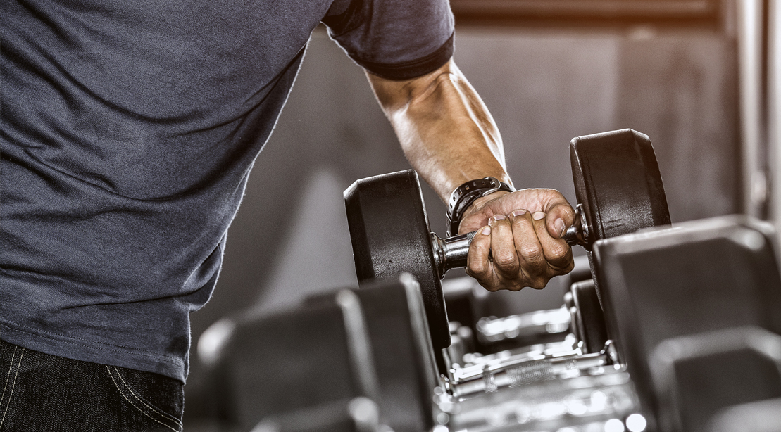 Homem musculoso segurando barra com pesos, ao lado de kettlebell e um aparelho de musculação high‑tech iluminado