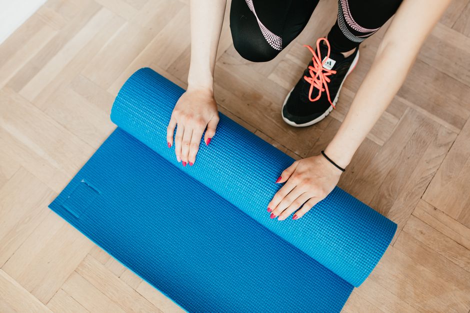 Mulher de 62 anos em roupa esportiva, segurando a parede enquanto eleva as pernas, com colchonete e bola de pilates ao fundo