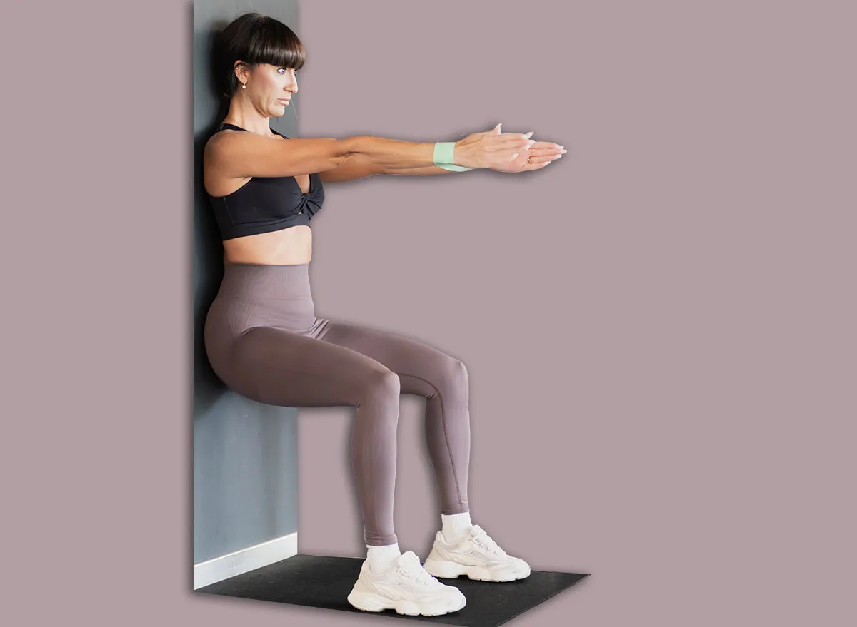 Mulher de meia-idade sentada em uma parede, com pernas estendidas e braços apoiados, demonstrando o exercício de wall sit
